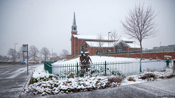 Chesterfield church snow The image shows a morning winter scene featuring the architecture of the Church of Jesus Christ of the Latter Day Saints in Chesterfield, Derbyshire, United Kingdom. Snow is falling gently and covers the church grounds, nearby trees, and a street that runs alongside the property. In the foreground, a statue is positioned behind a green metal fence, partially covered with snow. This is an urban landscape photograph that prominently displays the church, the snow-laden street, and surrounding trees, capturing the quiet atmosphere of Chesterfield during winter.
